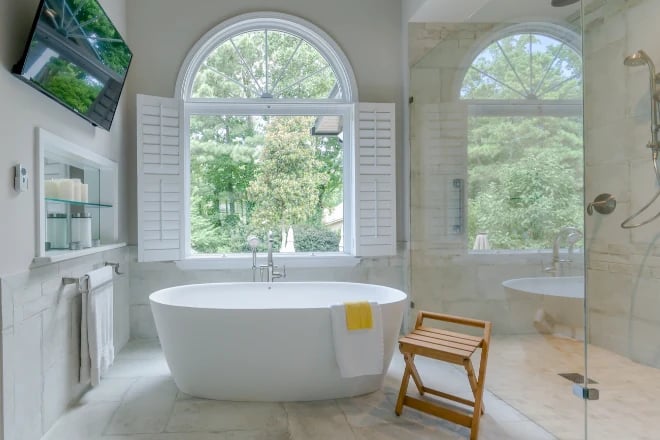 A freestanding tub in front of a window next to a small wooden stool